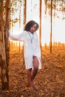 A woman stands barefoot in a serene forest setting during sunset, wearing a flowing white dress and a beaded necklace. The warm golden light creates long shadows among the tall, slender trees with peeling bark, enhancing the natural and peaceful atmosphere.