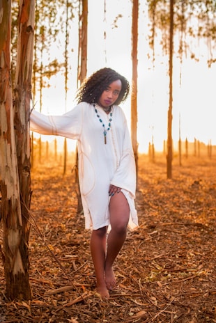 A woman stands barefoot in a serene forest setting during sunset, wearing a flowing white dress and a beaded necklace. The warm golden light creates long shadows among the tall, slender trees with peeling bark, enhancing the natural and peaceful atmosphere.