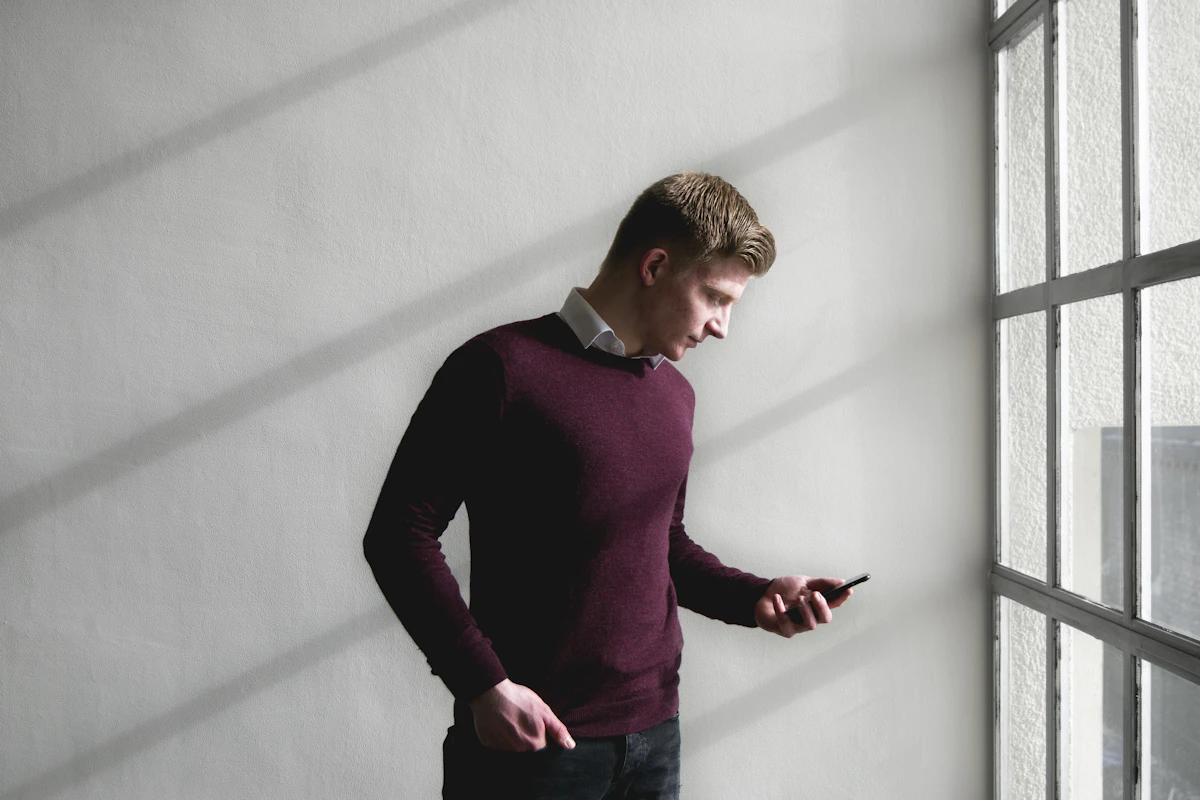 A man in a burgundy sweater checks his phone beside a tall paned window in a bare white room