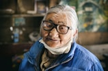 An elderly woman with long white hair tied back, wearing large black-framed glasses and a blue winter jacket. A white mask hangs below her chin. The background is blurred, featuring various elements that suggest an indoor setting, possibly within a home or rustic environment. The woman's gentle expression adds warmth to the scene, contrasting with the muted tones of the background.