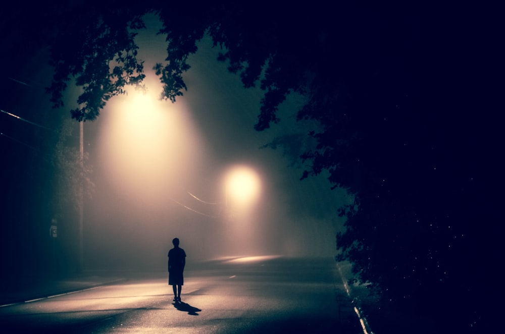 silhouette of person standing on concrete road with streetlights turned on during nighttime