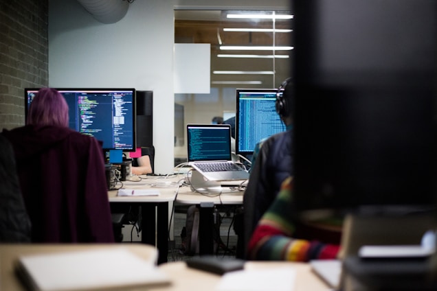 Several people are working at desks with computers displaying lines of code. The room has a modern office environment with overhead lighting and a glass partition. One person has purple hair, and another is wearing headphones.