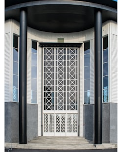 A symmetrical, modern building entrance with large black pillars and tall doors featuring geometric patterns. The exterior includes textured gray walls and large windows, creating an imposing architectural style.
