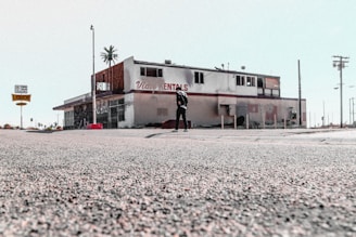 The image depicts an old, possibly abandoned rental building with a weathered appearance. The building has visible signs of wear and tear, missing parts, and graffiti. A person stands alone in front of the structure. The surrounding area appears desolate, with sparse traffic signs and distant palm trees.