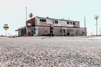 The image depicts an old, possibly abandoned rental building with a weathered appearance. The building has visible signs of wear and tear, missing parts, and graffiti. A person stands alone in front of the structure. The surrounding area appears desolate, with sparse traffic signs and distant palm trees.