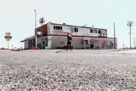 The image depicts an old, possibly abandoned rental building with a weathered appearance. The building has visible signs of wear and tear, missing parts, and graffiti. A person stands alone in front of the structure. The surrounding area appears desolate, with sparse traffic signs and distant palm trees.