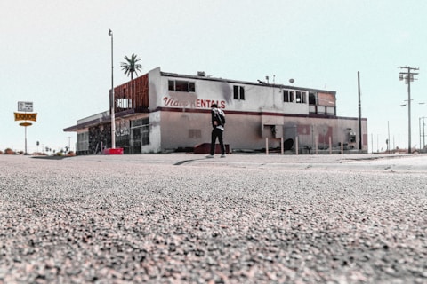 The image depicts an old, possibly abandoned rental building with a weathered appearance. The building has visible signs of wear and tear, missing parts, and graffiti. A person stands alone in front of the structure. The surrounding area appears desolate, with sparse traffic signs and distant palm trees.