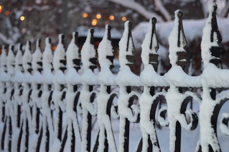An ornate black wrought iron fence is covered in a layer of white snow, creating a stark contrast. The fence features pointed tops and intricate designs typical of traditional ironwork. In the blurred background, there is a hint of warm yellow and orange lights, suggesting a cozy, possibly evening setting.