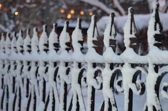 An ornate black wrought iron fence is covered in a layer of white snow, creating a stark contrast. The fence features pointed tops and intricate designs typical of traditional ironwork. In the blurred background, there is a hint of warm yellow and orange lights, suggesting a cozy, possibly evening setting.