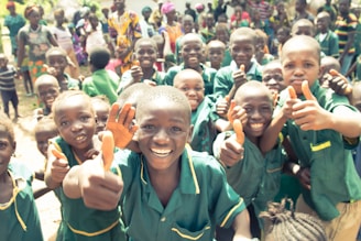 A group of cheerful children wearing green uniforms are giving thumbs up to the camera. They are outside, surrounded by other children and adults in the background, who are also smiling and seem to be enjoying themselves. The children are very close to each other, indicating a sense of camaraderie and joy.