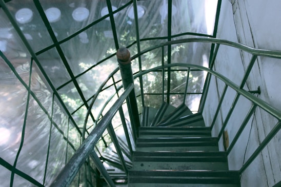 A spiral staircase with green metal railings, surrounded by glass walls that reflect the light and nearby trees. The staircase appears industrial and narrow, leading downward in a tight coil.