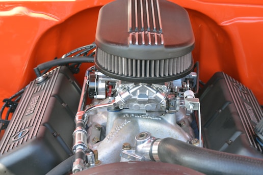 An engine with a prominent air filter on top, surrounded by various metallic components and fittings. The engine is set within a bright orange backdrop, highlighting its complex mechanical structure.