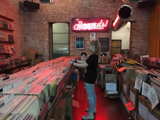 A young person with long hair stands amidst rows of vinyl records in a cozy record store with exposed brick walls. They are browsing through the records that are organized in crates arranged on long tables. A neon sign reading 'Records!' glows on the wall, casting a reddish light over the scene, while various posters and framed records decorate the space.