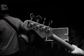 A black and white photograph capturing the close-up view of a guitar neck with tuning pegs. In the background, a musician wearing a hat and a t-shirt holds the guitar. An audience can be seen in dim lighting, seated on plastic chairs.