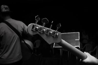 A black and white photograph capturing the close-up view of a guitar neck with tuning pegs. In the background, a musician wearing a hat and a t-shirt holds the guitar. An audience can be seen in dim lighting, seated on plastic chairs.