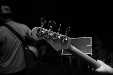 A black and white photograph capturing the close-up view of a guitar neck with tuning pegs. In the background, a musician wearing a hat and a t-shirt holds the guitar. An audience can be seen in dim lighting, seated on plastic chairs.