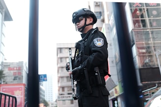 A security officer in protective gear stands in a city environment. The officer is wearing a black uniform, helmet, and sunglasses, holding a baton. The urban backdrop includes buildings and various signs with some visible text in a foreign language.