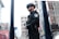 A security officer in protective gear stands in a city environment. The officer is wearing a black uniform, helmet, and sunglasses, holding a baton. The urban backdrop includes buildings and various signs with some visible text in a foreign language.