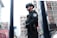 A security officer in protective gear stands in a city environment. The officer is wearing a black uniform, helmet, and sunglasses, holding a baton. The urban backdrop includes buildings and various signs with some visible text in a foreign language.