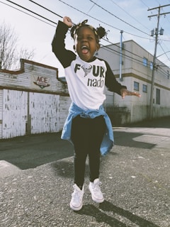 A joyful child is captured mid-jump on an outdoor street with a playful expression. The child wears a white shirt with a design featuring the word 'nado', a denim jacket tied around the waist, black pants, and white sneakers. Behind the child is a mural with the word 'ACE', next to some industrial-style buildings and power lines.