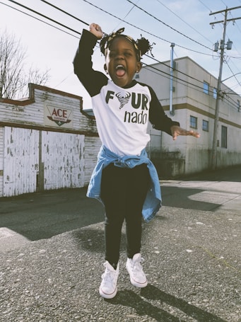 A joyful child is captured mid-jump on an outdoor street with a playful expression. The child wears a white shirt with a design featuring the word 'nado', a denim jacket tied around the waist, black pants, and white sneakers. Behind the child is a mural with the word 'ACE', next to some industrial-style buildings and power lines.