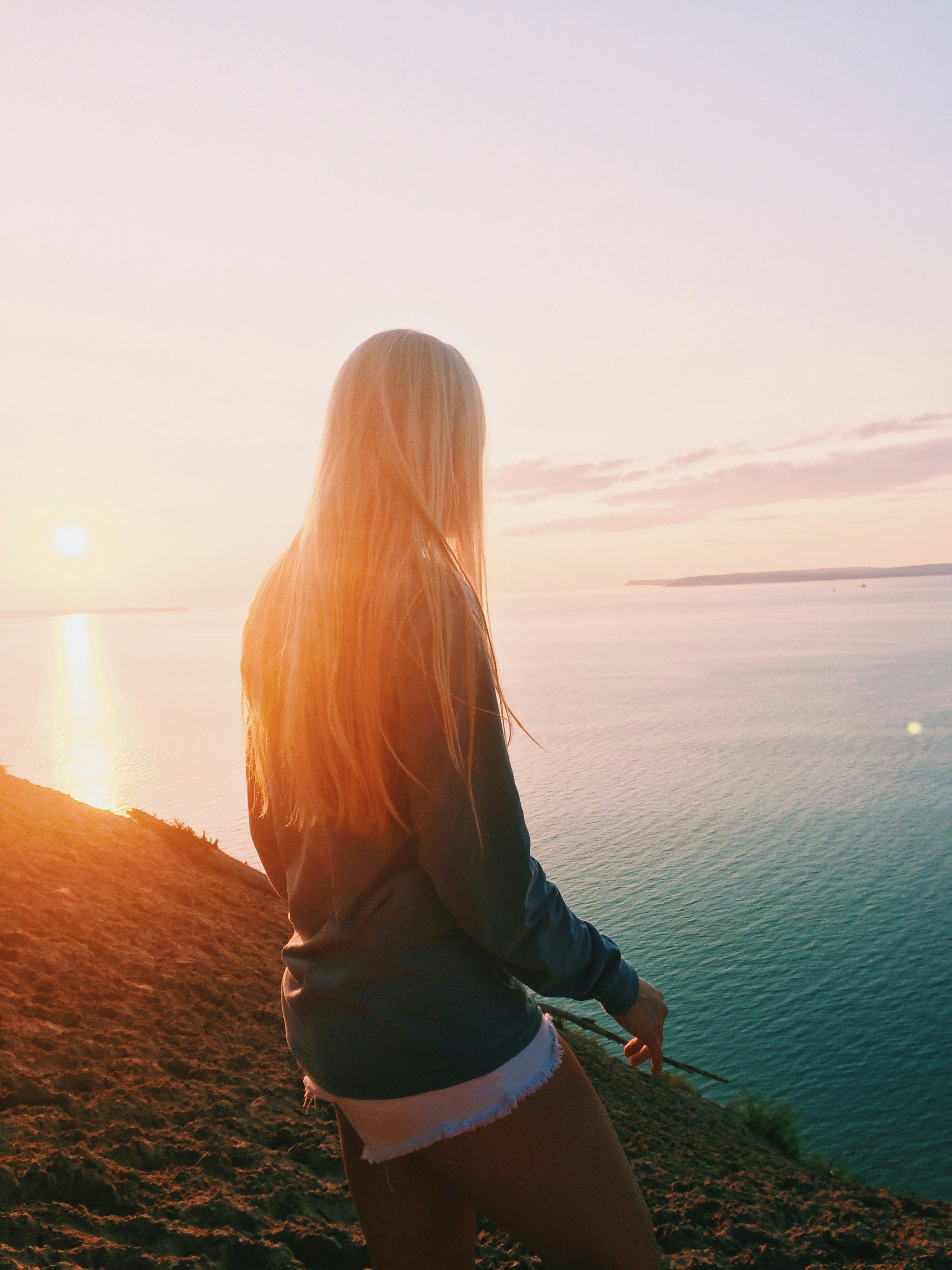 A woman with long, straight blonde hair stands on a cliff at sunset, showcasing a natural look ideal for evaluating Micro K-Tip extension lengths.