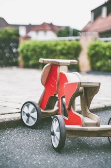 A wooden, red ride-on toy is placed on a paved pathway, with a background of blurry residential buildings and green hedges.