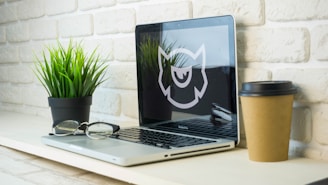 A workspace setup featuring a laptop with a stylized motif displayed on the screen, a pair of eyeglasses resting on the keyboard, a potted plant with vibrant green leaves, and a takeaway coffee cup placed on a light-colored shelf against a textured white brick wall.
