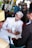 A man dressed in a formal white outfit is signing a document on a table. He is wearing a patterned headscarf and seated at an event with floral decorations in the background. Several people, some wearing traditional attire, are surrounding him. A microphone is placed on the table.