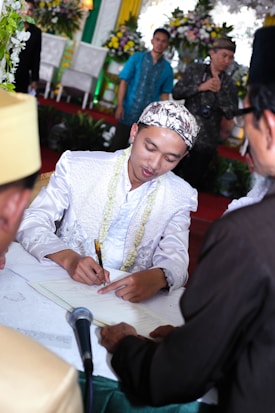 A man dressed in a formal white outfit is signing a document on a table. He is wearing a patterned headscarf and seated at an event with floral decorations in the background. Several people, some wearing traditional attire, are surrounding him. A microphone is placed on the table.