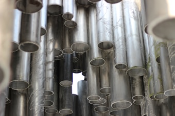 A collection of metallic pipes with textured surfaces and varying diameters is arranged closely together. Some pipes have a weathered appearance with subtle engravings or patterns, and the light filters through the gaps, creating a cool and industrial atmosphere.