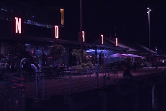 A nighttime scene with a building featuring prominently lit letters spelling 'NORTH' in red neon. People are sitting at tables outdoors under umbrellas, and there are plants and decorative lights along the railing. The atmosphere is lively with a modern aesthetic.