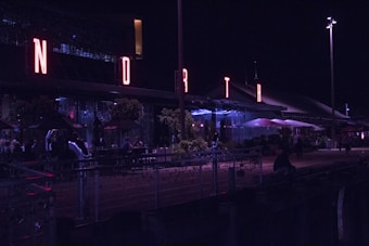 A nighttime scene with a building featuring prominently lit letters spelling 'NORTH' in red neon. People are sitting at tables outdoors under umbrellas, and there are plants and decorative lights along the railing. The atmosphere is lively with a modern aesthetic.