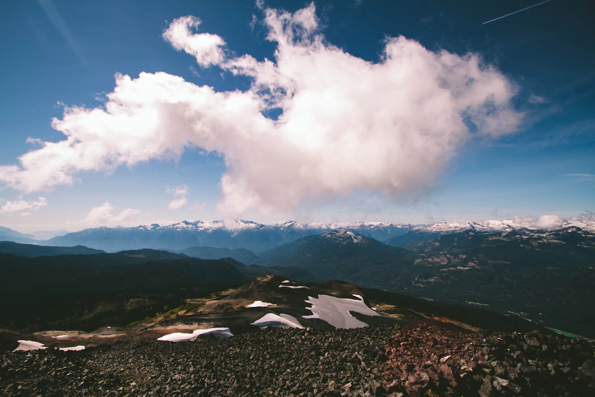 Rocky alpine ground with patches of snow under a big white cloud, looking across forested valleys to distant snowy peaks
