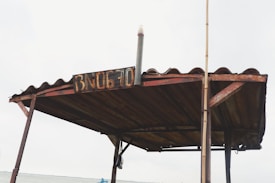 A rustic metal roof structure with a weathered sign displaying the text 'BN0670'. The roof is supported by metal poles and has a corrugated design, evoking a sense of aged industrial utility. A long vertical pole is attached to one side of the structure.