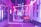 A narrow urban alley illuminated with vibrant pink and purple neon lights. The lights create a surreal and atmospheric ambiance. There is a Spanish flag hanging on one of the elements in the foreground, and a sign with the word 'ROXY' is visible on the side of a building. Blurred figures suggest movement or a slow shutter speed.