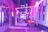 A narrow urban alley illuminated with vibrant pink and purple neon lights. The lights create a surreal and atmospheric ambiance. There is a Spanish flag hanging on one of the elements in the foreground, and a sign with the word 'ROXY' is visible on the side of a building. Blurred figures suggest movement or a slow shutter speed.