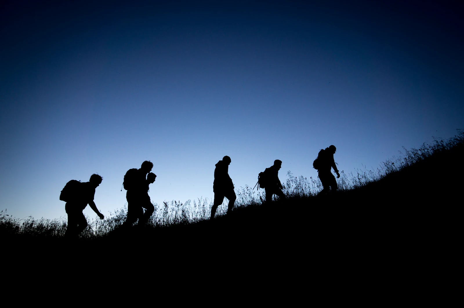 A group of people with backpacks hiking up a grassy hill in silhouette