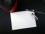 A blank white sheet of paper lies on a dark surface surrounded by a pen and a small container. In the upper left corner, part of a red and black speaker is visible.