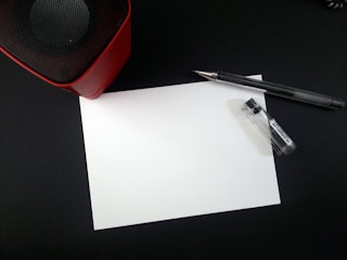 A blank white sheet of paper lies on a dark surface surrounded by a pen and a small container. In the upper left corner, part of a red and black speaker is visible.