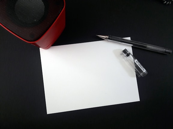 A blank white sheet of paper lies on a dark surface surrounded by a pen and a small container. In the upper left corner, part of a red and black speaker is visible.