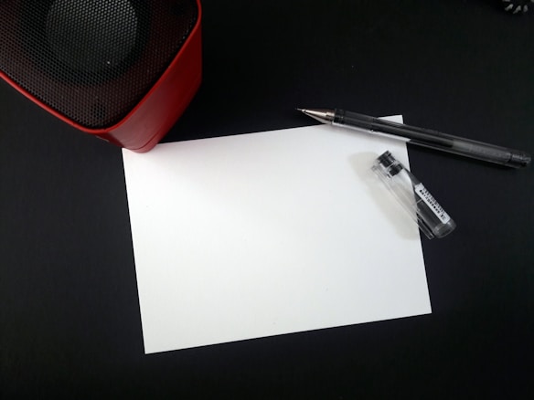 A blank white sheet of paper lies on a dark surface surrounded by a pen and a small container. In the upper left corner, part of a red and black speaker is visible.