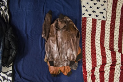 A brown leather jacket is laid out flat on a dark blue fabric with part of a folded American flag visible on the right. On the left, there is a glimpse of another dark-colored item, possibly another jacket or blanket.