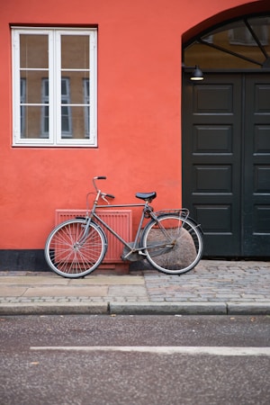 A vintage bicycle is parked against a vibrant orange wall. The wall features a white-framed window and a large, dark green door. The scene is set on a cobblestone path next to a paved road.