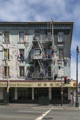 A multi-story building with murals and intricate artwork on the facade. The lower part houses a Hong Kong restaurant with bilingual signage. A fire escape ladder runs vertically, and various people are depicted in the murals. Bright sunlight casts shadows on the building, and pedestrians and street elements like traffic lights are present in the foreground.