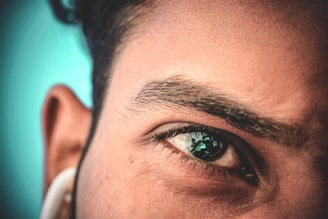 A close-up view of a person's face, highlighting their eye, eyebrow, forehead, and ear. The eye reflects an outdoor scene with apparent greenery. The image is detailed enough to show the texture of the skin and individual hairs of the eyebrow.