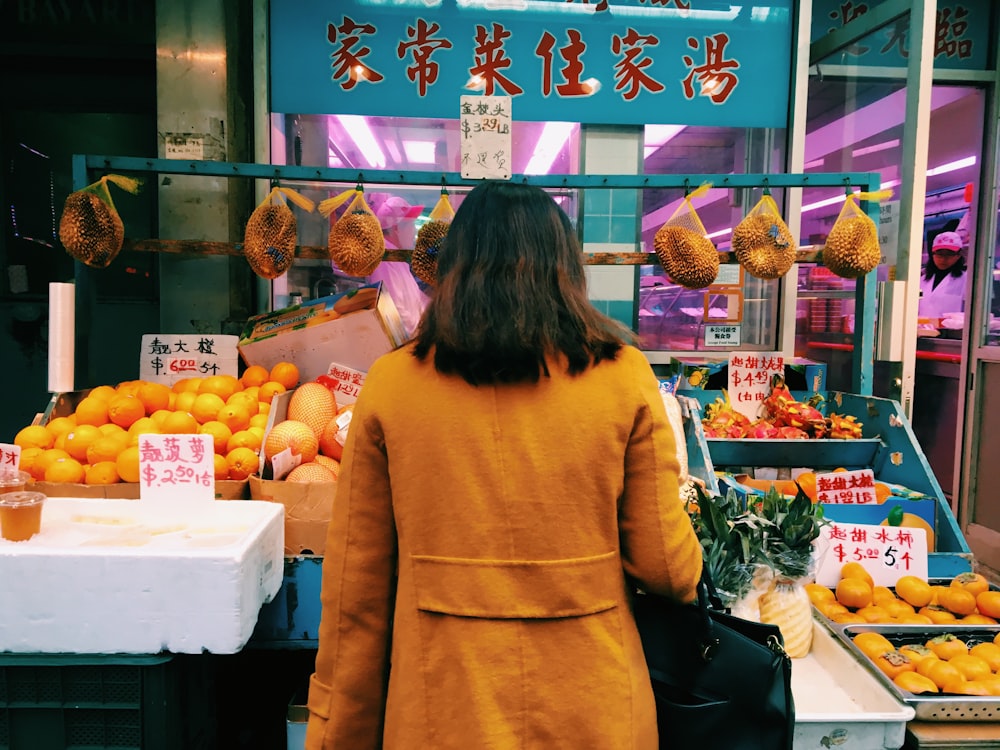 standing woman wearing brown coat toward display fruits