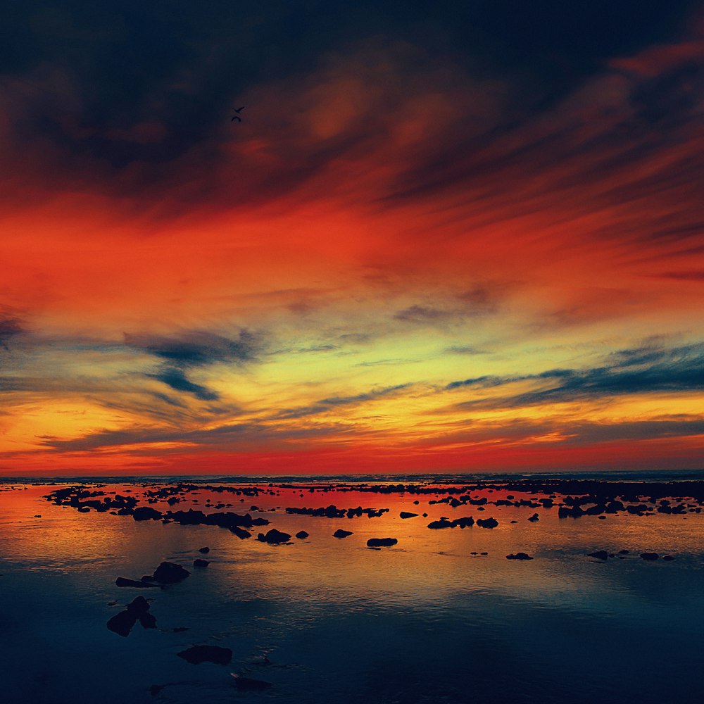 silhouette of rocks on body of water under red and blue clouds