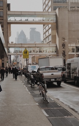 An urban street scene with pedestrians walking along a sidewalk, bicycles parked along the side, and vehicles including a truck and vans on the road. Large buildings surround the street, and elevated pedestrian walkways connect the structures, with people visible inside. The cityscape includes skyscrapers in the background, partially obscured by haze or fog.