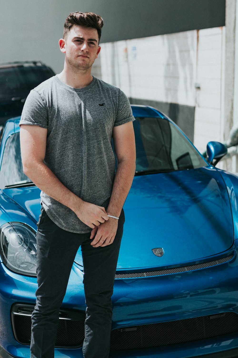 man wearing grey t-shirt and black pants standing in front of blue Porsche vehcile